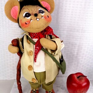 May include: A plush mouse doll wearing a tan jacket, olive green pants, a red bandana, and a green hat. The mouse holds a brown walking stick and stands next to a large, shiny red apple. The doll features large ears with pink centers and painted facial details.