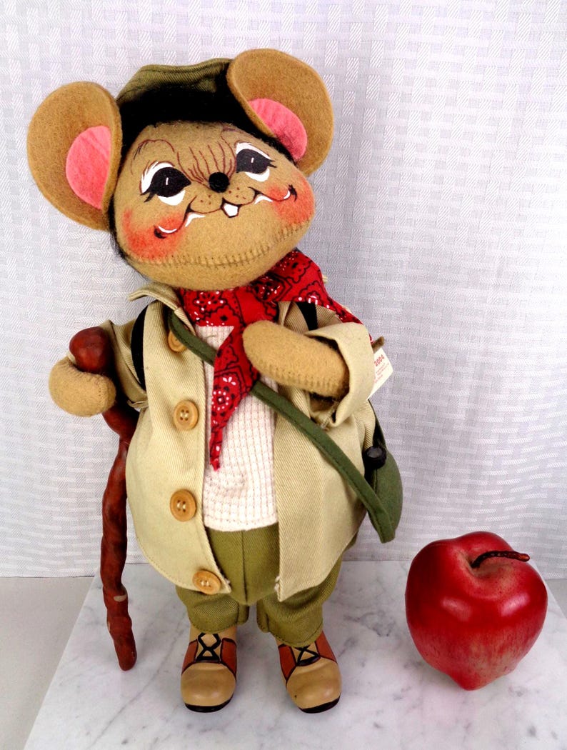 May include: A plush mouse doll wearing a tan jacket, olive green pants, a red bandana, and a green hat. The mouse holds a brown walking stick and stands next to a large, shiny red apple. The doll features large ears with pink centers and painted facial details.