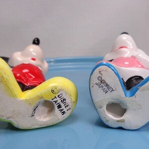 May include: Two vintage Mickey Mouse figurines, viewed from the bottom, with yellow, red, white, and black details. One is marked "Disney Taiwan" and the other "Disney Japan". The figurines are on a light blue surface.