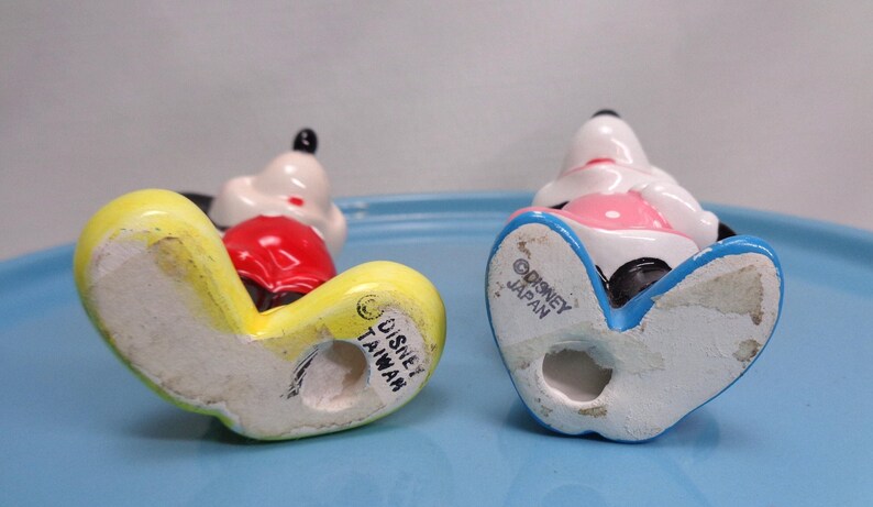May include: Two vintage Mickey Mouse figurines, viewed from the bottom, with yellow, red, white, and black details. One is marked "Disney Taiwan" and the other "Disney Japan". The figurines are on a light blue surface.