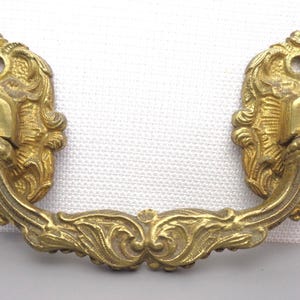 May include: Ornate brass drawer pull with a curved handle and decorative floral and scrollwork details. The pull features a rectangular backplate with a keyhole and a detailed, textured design. A vintage-style hardware piece.