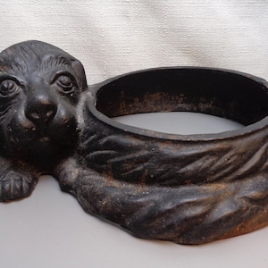 May include: A cast iron dog planter with a round opening for planting. The dog is lying down with its head resting on the planter.