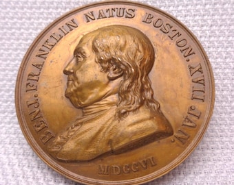 May include: A circular bronze-toned commemorative medal featuring a profile relief of Benjamin Franklin facing left. The inscription reads BENJ. FRANKLIN NATUS BOSTON XVII JAN. MDCCVI along the raised border of the metallic coin-like object.