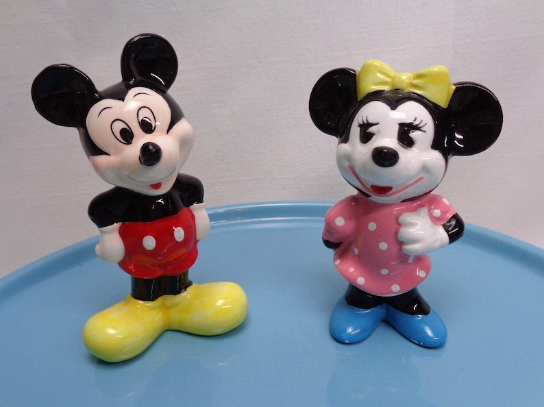May include: Two ceramic figurines of Mickey and Minnie Mouse. Mickey is wearing a black top, red shorts with white buttons, and yellow shoes. Minnie is in a pink polka-dot dress, blue shoes, and a yellow bow.