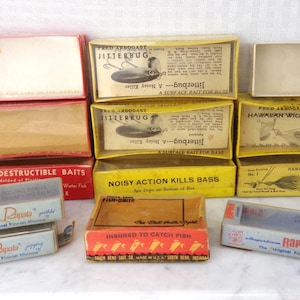May include: A collection of vintage fishing lure boxes in various colors, including red, yellow, and blue. The boxes feature printed text and graphics, with brands like Jitterbug, Hawaiian Wiggler, and Rapala. Some boxes are open, revealing the contents.