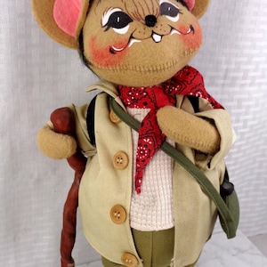 May include: A plush mouse doll wearing a tan jacket with three buttons, a white textured shirt, olive green pants, and a red bandana with a black pattern. The mouse has large ears with pink centers, a brown walking stick, and brown hiking boots.