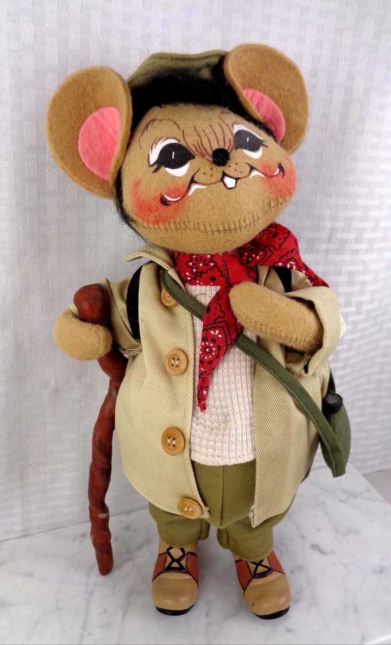 May include: A plush mouse doll wearing a tan jacket with three buttons, a white textured shirt, olive green pants, and a red bandana with a black pattern. The mouse has large ears with pink centers, a brown walking stick, and brown hiking boots.