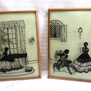 May include: Two framed vintage silhouette art pieces. Each features a black silhouette scene on a light green background, set within a copper-colored frame. One depicts a woman at a table, the other a couple.