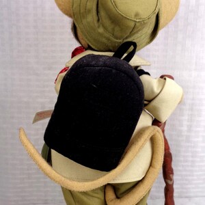 May include: A plush mouse doll dressed in a tan safari outfit, including a matching bucket hat and jacket. The mouse wears a black backpack and carries a brown walking stick. A long, curved, tan tail extends from the back of the doll's pants.