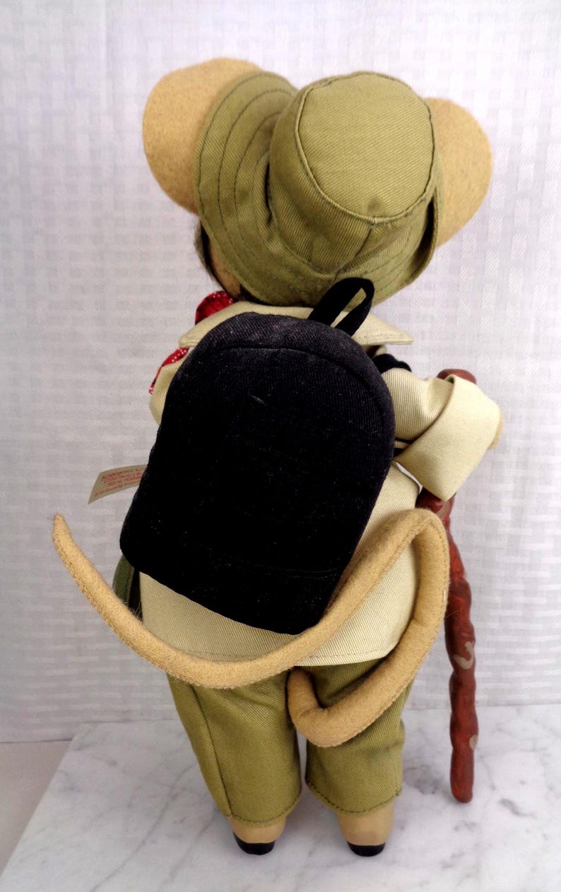 May include: A plush mouse doll dressed in a tan safari outfit, including a matching bucket hat and jacket. The mouse wears a black backpack and carries a brown walking stick. A long, curved, tan tail extends from the back of the doll's pants.