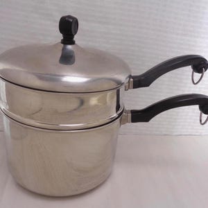 May include: A stainless steel double boiler with a lid and two black handles. The lid has a black knob. The handles have metal rings at the ends. The pot is designed for steaming or melting ingredients.