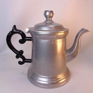 May include: A silver teapot with a black handle and a lid. The teapot has a round body and a spout on the side. The teapot is made of metal and has a shiny finish.
