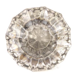 May include: A clear, faceted glass doorknob with a circular shape. The knob has a detailed, textured surface with a central design. The glass reflects light, creating highlights and shadows. The doorknob is isolated against a white background.