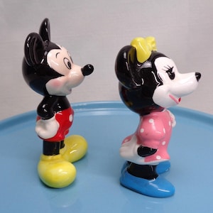 May include: Two ceramic figurines of Mickey and Minnie Mouse. Mickey is in red shorts, yellow shoes, and black ears. Minnie wears a pink dress with white polka dots, a yellow bow, and blue shoes. Both have black and white faces.
