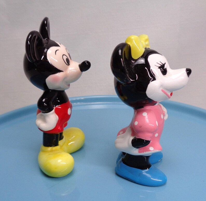 May include: Two ceramic figurines of Mickey and Minnie Mouse. Mickey is in red shorts, yellow shoes, and black ears. Minnie wears a pink dress with white polka dots, a yellow bow, and blue shoes. Both have black and white faces.