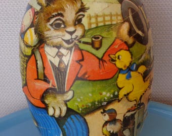 May include: A vintage Easter egg shaped like a rabbit wearing a red jacket, blue pants, and a tie. The rabbit is holding a pipe and carrying a basket of eggs. A yellow chick with a blue ribbon is standing next to the rabbit. The egg is decorated with a green, brown, and white background.