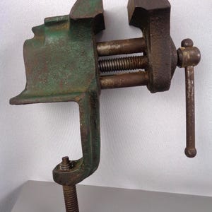 May include: An antique bench vise with a green and brown finish. The vise has a screw mechanism for clamping and a handle for tightening. The tool is designed to be clamped to a workbench and used for holding objects securely during woodworking or metalworking tasks.