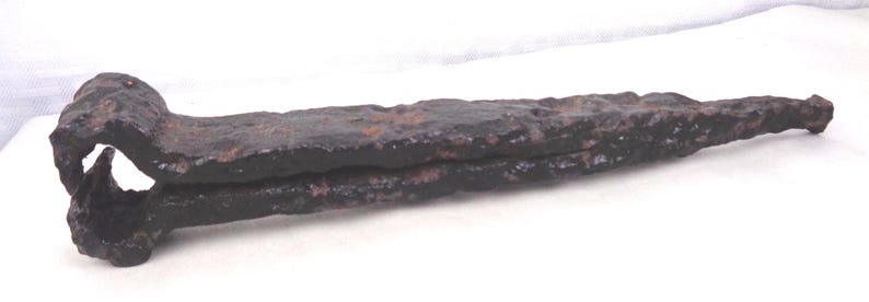May include: A large, antique iron nail with a rounded head and a long, tapering shank. The nail is heavily rusted, displaying a dark brown color. The head has a distinctive curved shape, and the overall design suggests a historical or vintage item.