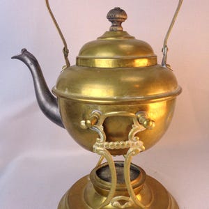 May include: Vintage brass tea kettle with a black wooden handle and a decorative stand. The kettle features a rounded body, a curved spout, and a lid with a small finial. The stand has an ornate design and a circular base.