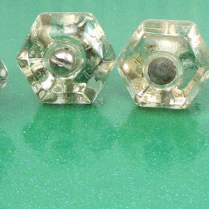 Peut inclure: Six boutons de meuble en verre clair et facetté, de forme hexagonale. Chaque bouton a une vis argentée et un centre gris foncé. Les boutons sont disposés sur une surface verte.