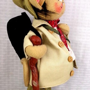 May include: A plush mouse figurine dressed as a hiker, wearing a tan jacket with buttons, olive green pants, and a matching hat. The mouse carries a black backpack, a red patterned neckerchief, and holds a textured brown walking stick.