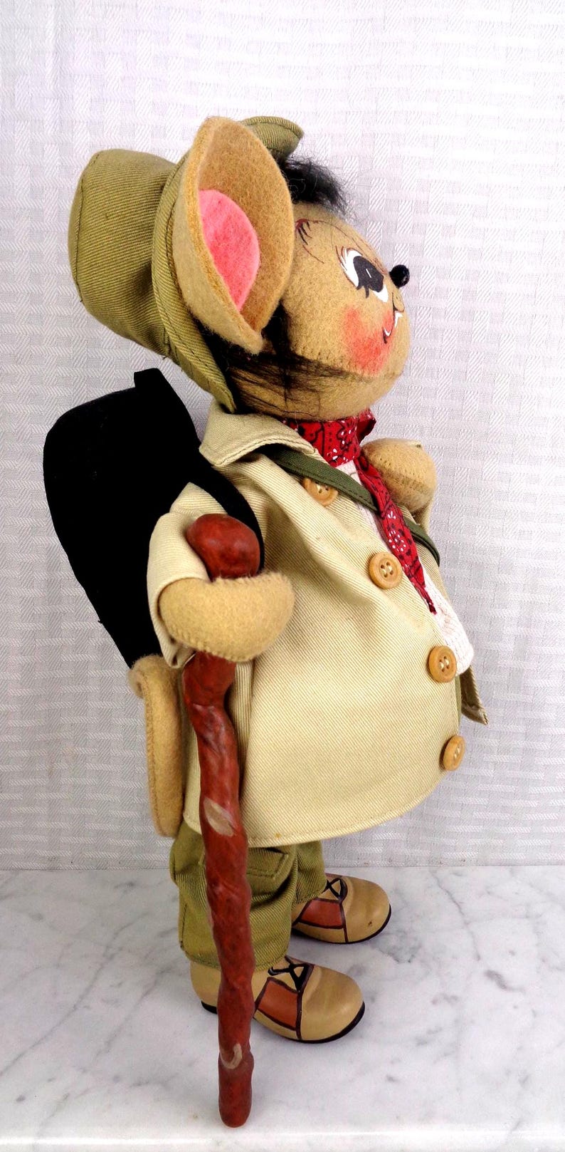 May include: A plush mouse figurine dressed as a hiker, wearing a tan jacket with buttons, olive green pants, and a matching hat. The mouse carries a black backpack, a red patterned neckerchief, and holds a textured brown walking stick.
