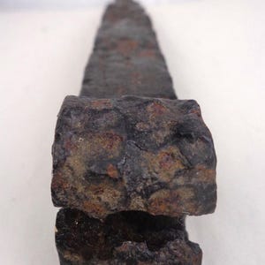 May include: A weathered, dark gray metal object with a rectangular base and a tapered, slightly rounded top. The surface shows signs of rust and wear, suggesting age. The object appears to be a tool or a part of a larger structure.