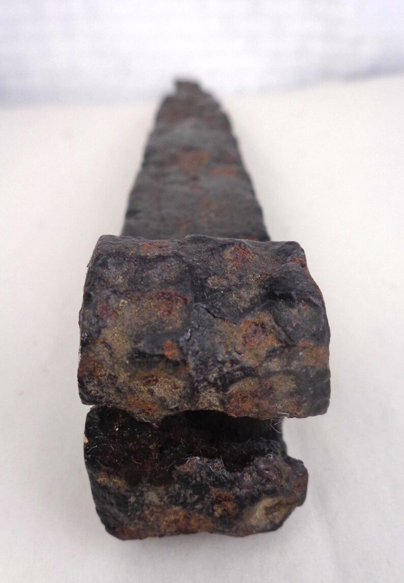 May include: A weathered, dark gray metal object with a rectangular base and a tapered, slightly rounded top. The surface shows signs of rust and wear, suggesting age. The object appears to be a tool or a part of a larger structure.