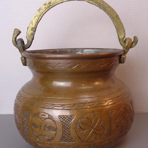 May include: A copper pot with a brass handle. The pot has an ornate design with a floral pattern. The pot is about 10 inches tall.