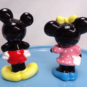 May include: A pair of ceramic figurines depicting Mickey and Minnie Mouse. Mickey is in red shorts, yellow shoes, and white gloves. Minnie wears a pink dress with white polka dots, a yellow bow, and blue shoes.