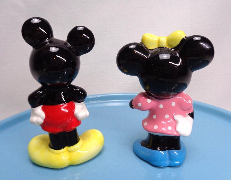 May include: A pair of ceramic figurines depicting Mickey and Minnie Mouse. Mickey is in red shorts, yellow shoes, and white gloves. Minnie wears a pink dress with white polka dots, a yellow bow, and blue shoes.
