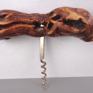 May include: A rustic corkscrew with a natural wood handle. The handle is a dark brown color with lighter wood grain patterns and a silver metal screw. The corkscrew is designed for removing corks from wine bottles.