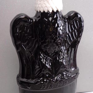 May include: A black glass eagle-shaped bottle with a white head. The bottle has detailed wing and feather designs. The base of the bottle has the year 1776. A decorative item.
