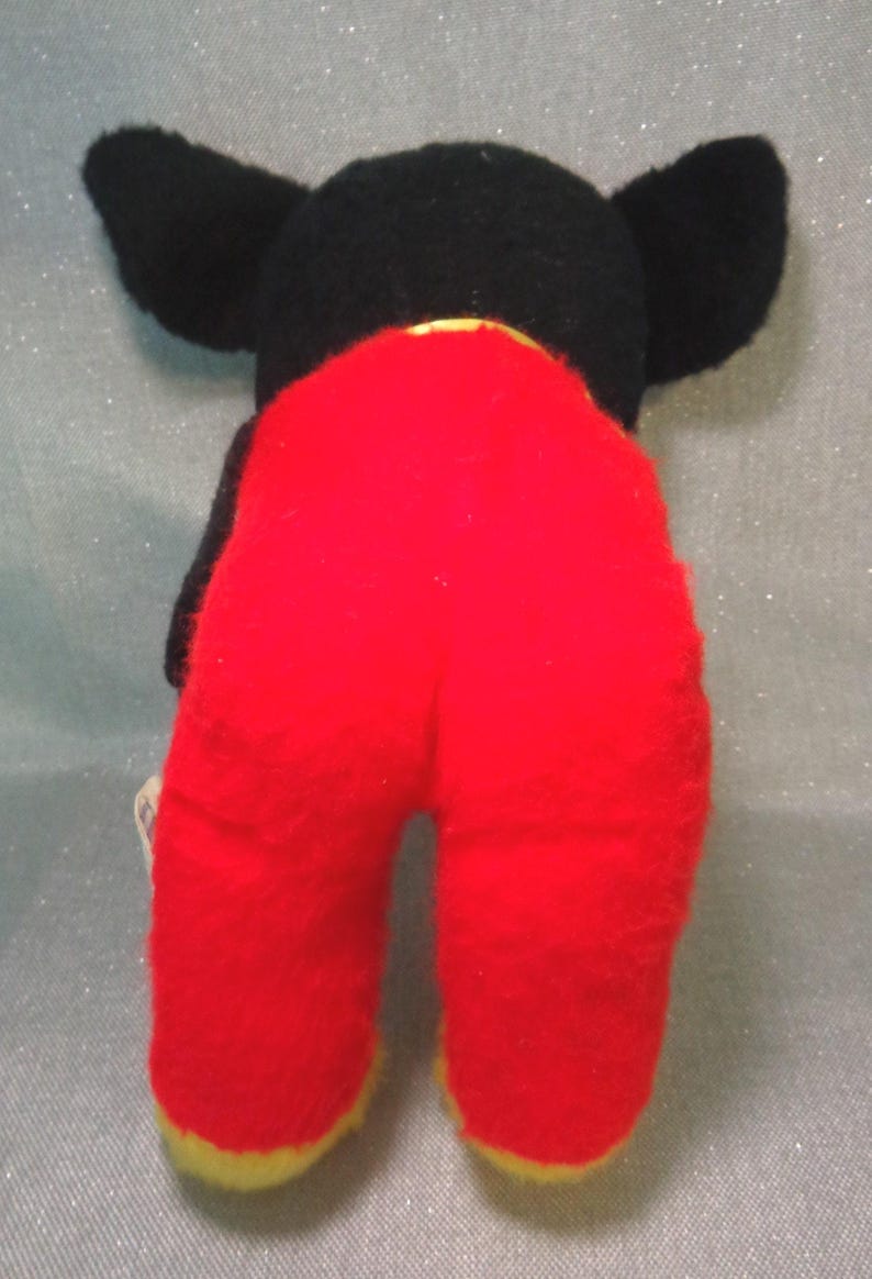 Puede incluir: Juguete de peluche negro y rojo con orejas grandes. El juguete lleva pantalones rojos con ribetes amarillos.