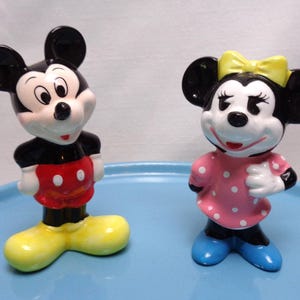 May include: Two ceramic figurines of Mickey and Minnie Mouse. Mickey is in a black top, red shorts with white buttons, and yellow shoes. Minnie is in a pink polka-dot dress, yellow bow, and blue shoes.