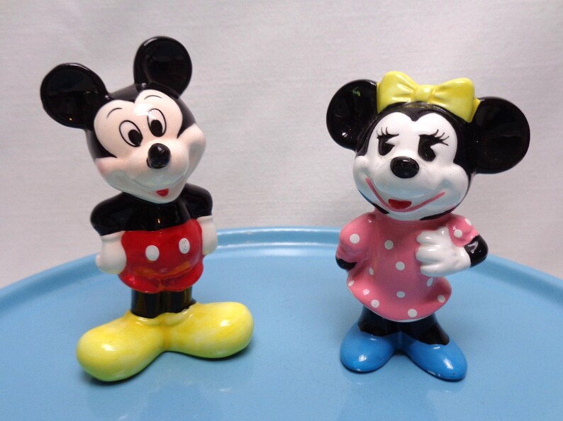 May include: Two ceramic figurines of Mickey and Minnie Mouse. Mickey is in a black top, red shorts with white buttons, and yellow shoes. Minnie is in a pink polka-dot dress, yellow bow, and blue shoes.