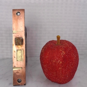 May include: A copper-toned metal door latch with rectangular and circular openings, next to a red decorative apple with a textured surface and a yellow stem. The latch is likely vintage.