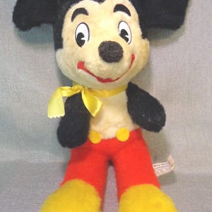 Puede incluir: Un juguete de peluche vintage de Mickey Mouse con orejas negras, un lazo amarillo, pantalones rojos y zapatos amarillos. El juguete tiene una cara blanca con ojos negros y una nariz negra. El juguete lleva una pajarita amarilla y tiene una etiqueta roja y amarilla que dice "Walt Disney Productions".