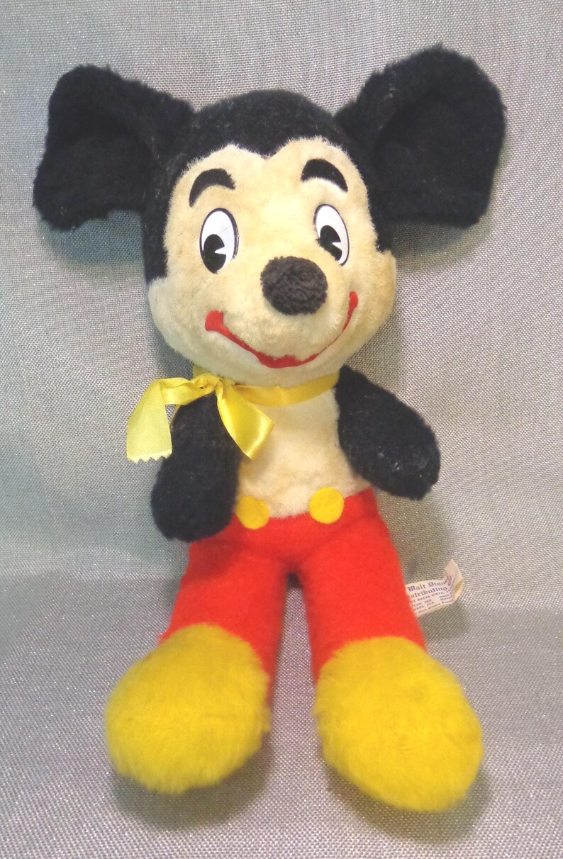 Puede incluir: Un juguete de peluche vintage de Mickey Mouse con orejas negras, un lazo amarillo, pantalones rojos y zapatos amarillos. El juguete tiene una cara blanca con ojos negros y una nariz negra. El juguete lleva una pajarita amarilla y tiene una etiqueta roja y amarilla que dice "Walt Disney Productions".
