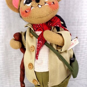 May include: A plush mouse doll wearing a tan jacket, olive green pants, and a red bandana. The mouse has large ears with pink centers, a dark green hat, and carries a brown walking stick and a green shoulder bag. A tag reads Annalee 2004.