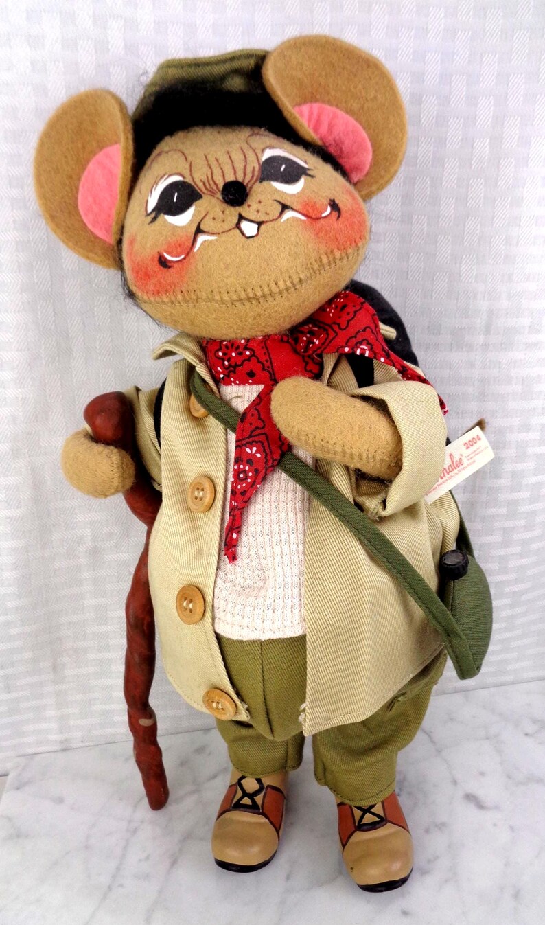 May include: A plush mouse doll wearing a tan jacket, olive green pants, and a red bandana. The mouse has large ears with pink centers, a dark green hat, and carries a brown walking stick and a green shoulder bag. A tag reads Annalee 2004.
