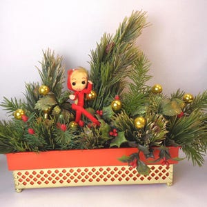 May include: A red and white Christmas elf doll sits in a red planter with green pine branches, gold ornaments, and red berries.