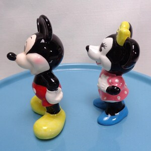 May include: A pair of ceramic figurines depicting Mickey and Minnie Mouse. Mickey is in red shorts, yellow shoes, and black ears. Minnie wears a pink polka-dot dress, blue shoes, and a yellow bow. Both have white faces and black features.