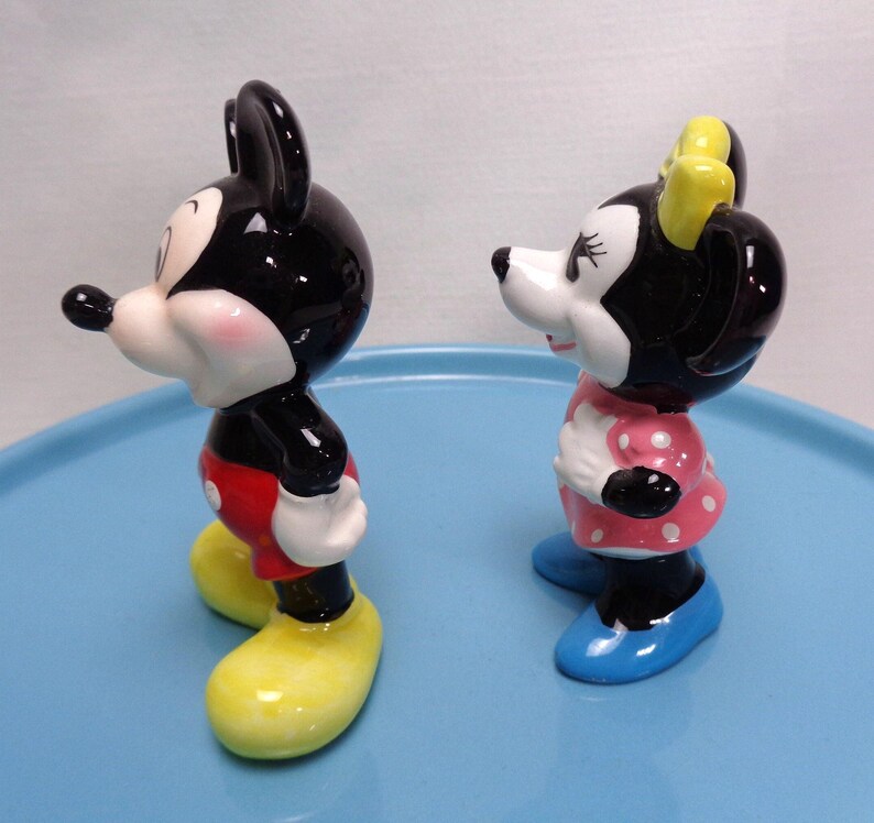 May include: A pair of ceramic figurines depicting Mickey and Minnie Mouse. Mickey is in red shorts, yellow shoes, and black ears. Minnie wears a pink polka-dot dress, blue shoes, and a yellow bow. Both have white faces and black features.