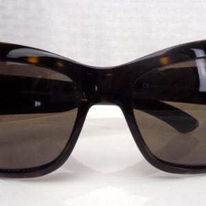 May include: Dark brown sunglasses with a tortoiseshell pattern on the frames. The lenses are a dark brown color, and the sunglasses have a curved, oversized design. The sunglasses are resting on a white surface.