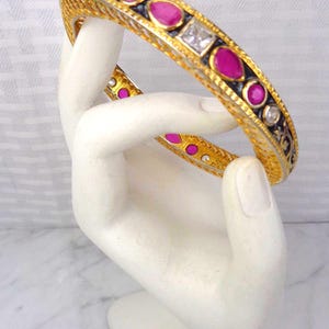 May include: Gold-toned bangle bracelet with a black and gold design, featuring oval and square pink gemstones and clear stones. The bracelet is displayed on a white hand-shaped sculpture.