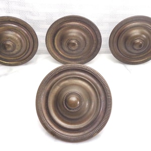 May include: A collection of six antique brass cabinet knobs. The knobs are circular with a central raised button and concentric rings. The outer edge of each knob has a decorative, ridged pattern. The knobs are arranged on a white surface.