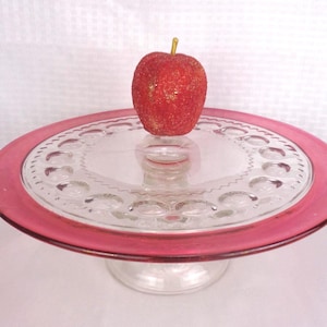 May include: A clear glass cake stand with a ruby red rim and a decorative pattern. A red apple with gold glitter sits on top. The stand is a vintage piece, perfect for displaying desserts.
