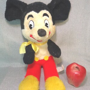 Puede incluir: Un juguete de peluche vintage de Mickey Mouse con orejas negras, una pajarita amarilla, pantalones rojos y zapatos amarillos. El juguete est&aacute; sentado en una superficie gris junto a una manzana roja.