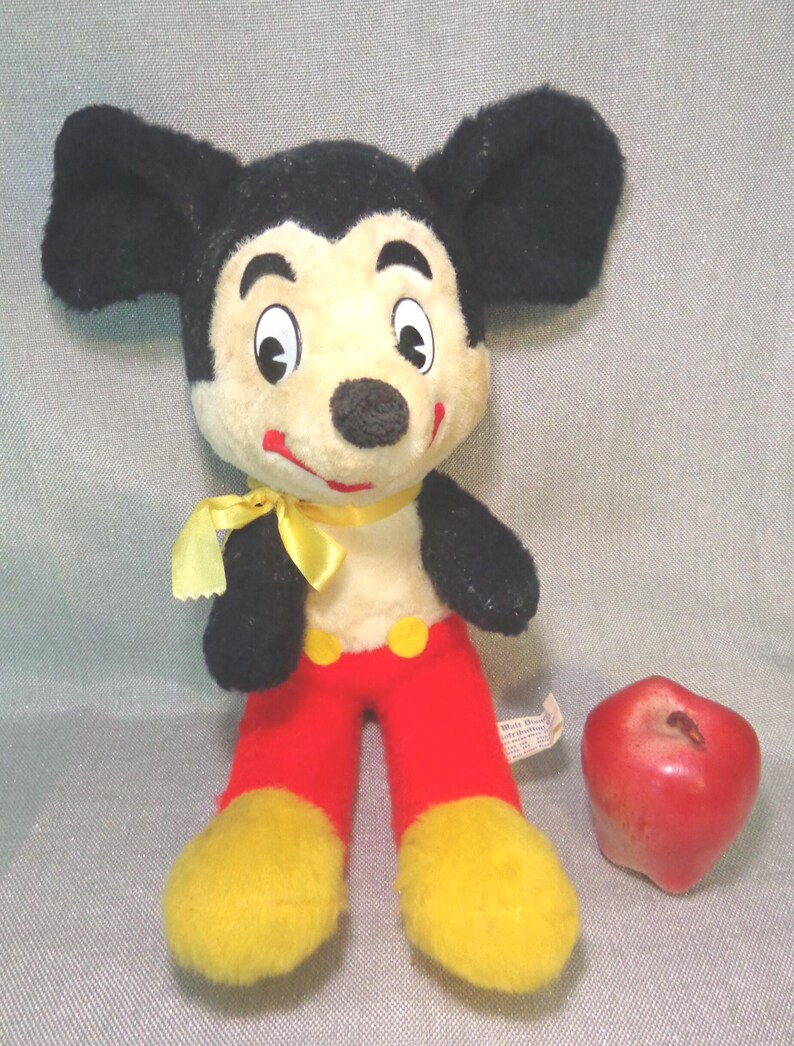Puede incluir: Un juguete de peluche vintage de Mickey Mouse con orejas negras, una pajarita amarilla, pantalones rojos y zapatos amarillos. El juguete est&aacute; sentado en una superficie gris junto a una manzana roja.